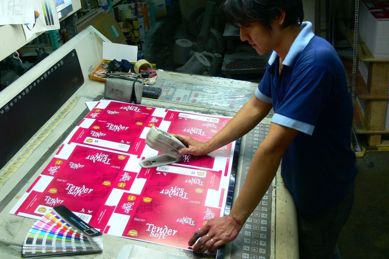A FirstPack operator reading a printed sheet with a handheld spectrophotometer — X-Rite unit and Pantone swatch book on the colour desk